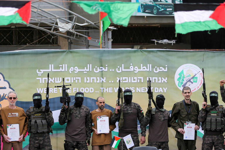 A imagem mostra um grupo de homens em trajes militares， alguns com máscaras， segurando armas. Entre eles， três reféns seguram documentos. Eles estão em frente a um fundo com bandeiras palestinas e um mural que contém texto em árabe. Os homens estão em poses de celebração， levantando os braços e as armas.