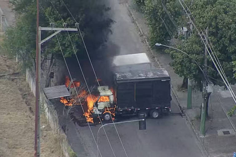 A imagem mostra um caminhão pegando fogo em uma rua. O veículo está envolto em chamas e fumaça preta se eleva em direção ao céu. Ao fundo， há árvores e postes de eletricidade， além de uma rua pavimentada.