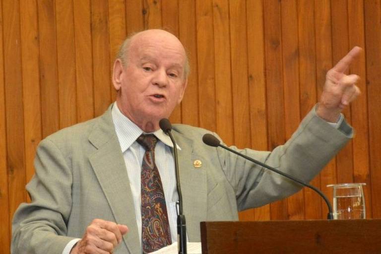 Um homem de terno claro e gravata， em pé， faz um discurso em uma câmara municipal. Ele está gesticulando com a mão direita levantada e parece estar enfatizando um ponto. Ao fundo， há uma parede de madeira e uma mesa com um copo de água. O homem tem cabelo claro e está falando em um microfone.