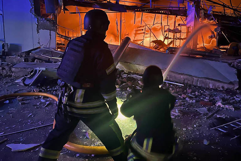 Em cenário noturno， dois bombeiros jogam água em incêndio numa ruína de edifício