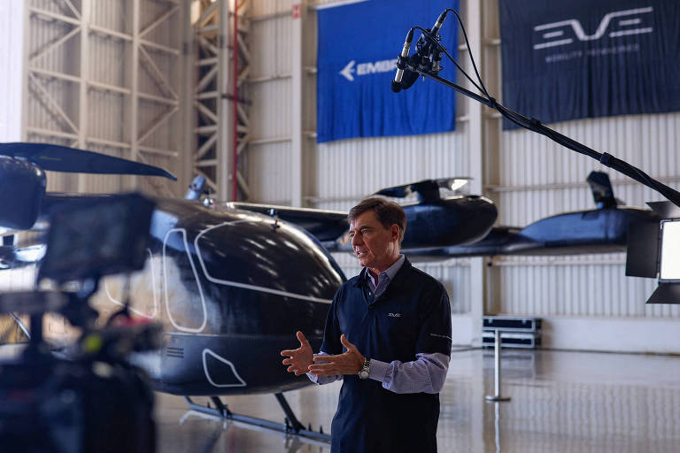 A imagem mostra um homem em um hangar， falando para uma câmera. Ao fundo， há um helicóptero preto e banners com os logotipos de empresas. O ambiente é bem iluminado e espaçoso.
