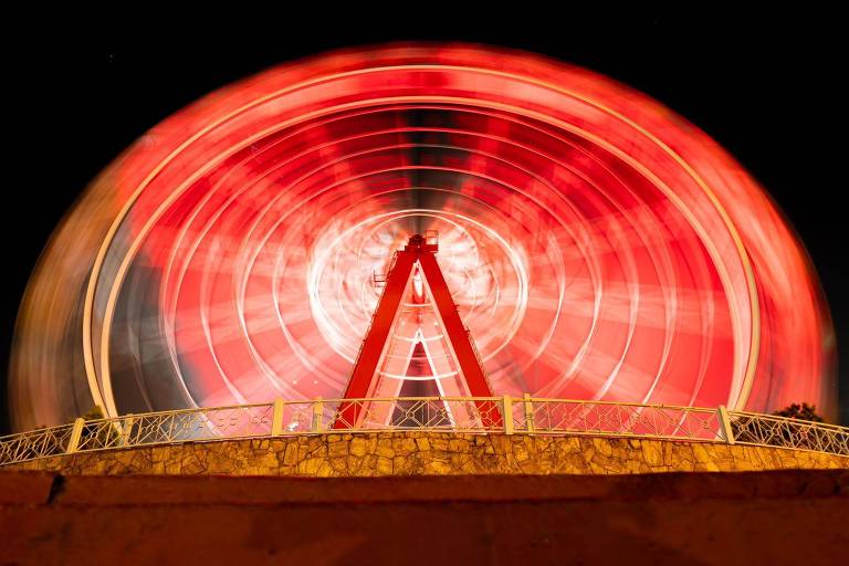 A imagem mostra uma roda gigante em movimento, com luzes vermelhas e brancas criando um efeito de desfoque. A estrutura da roda é visível na parte superior, enquanto a base é cercada por uma grade branca e um muro de pedra. O fundo é escuro, indicando que a foto foi tirada à noite.