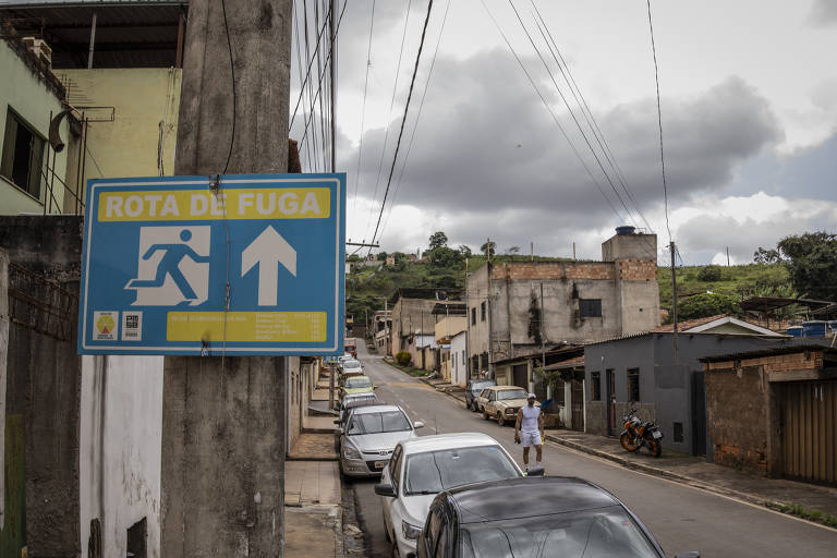 A imagem mostra uma rua em uma área urbana, com casas de diferentes tamanhos e condições. À esquerda, há um sinal azul com a inscrição 'ROTA DE FUGA' e uma seta apontando para cima, indicando a direção da rota. O céu está nublado, e há vários carros estacionados ao longo da rua. Uma pessoa vestindo roupas brancas caminha pela calçada.