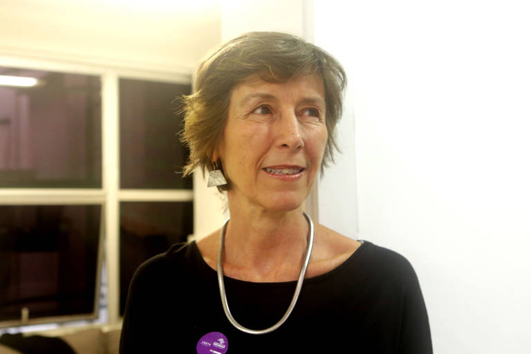 A imagem mostra uma mulher com cabelo curto e bagunçado， usando uma blusa preta e um colar prateado. Ela está em um ambiente interno， com janelas ao fundo. A mulher parece estar sorrindo levemente e tem um adesivo roxo visível em seu peito.