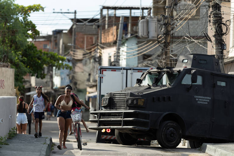 A imagem mostra uma rua em um bairro urbano， onde um veículo blindado da polícia está estacionado. Algumas pessoas estão caminhando pela calçada， incluindo uma mulher que anda de bicicleta. Ao fundo， há casas e fios elétricos visíveis， caracterizando um ambiente de comunidade. A cena sugere uma presença policial significativa na área.