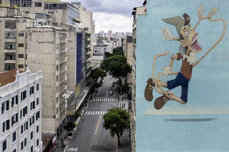 A imagem mostra um mural colorido de um personagem animado em uma parede de um edifício. O personagem está pulando e sorrindo, com um chapéu e roupas de cowboy. Ao fundo, há uma vista de uma rua urbana com prédios e árvores, e a rua está vazia. O céu está nublado.