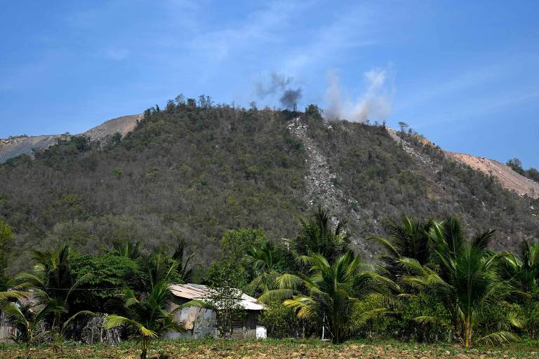 A imagem mostra uma montanha com vegetação escassa no topo, onde há fumaça saindo, indicando possível atividade de queimada. Na parte inferior, há uma casa simples cercada por palmeiras, sob um céu azul claro.