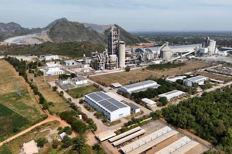 A imagem mostra uma vista aérea de uma fábrica localizada em uma área rural. A estrutura principal da fábrica é alta e possui várias chaminés. Ao redor, há edifícios menores e áreas verdes, com montanhas ao fundo. O céu está claro e a paisagem é predominantemente plana, com algumas áreas cultivadas visíveis.