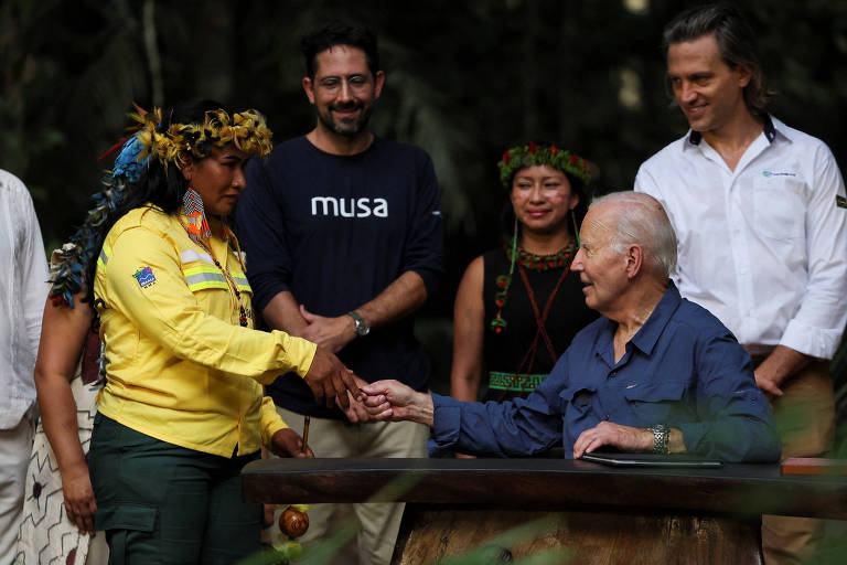 A imagem mostra um grupo de pessoas em uma cerimônia ao ar livre. Uma mulher com uma blusa amarela e adornos na cabeça está apertando a mão de um homem idoso， que está sentado à mesa. Ao fundo， há outras pessoas observando， incluindo um homem com uma camiseta preta e duas mulheres com trajes tradicionais. A cena parece ser de um evento significativo， possivelmente relacionado a questões ambientais. 