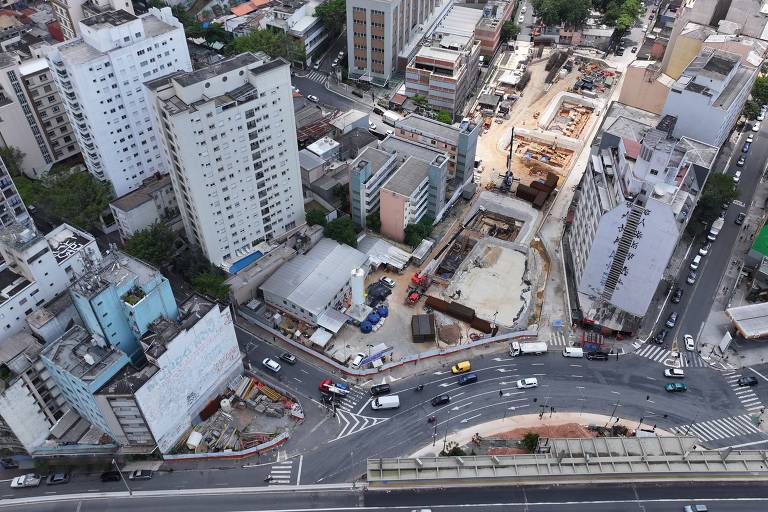 Imagem aérea de uma área urbana， mostrando diversos edifícios de diferentes alturas， ruas e veículos em movimento. Há uma construção em andamento visível no centro da imagem， cercada por outros prédios e áreas pavimentadas. A cena é predominantemente urbana， com uma mistura de estruturas residenciais e comerciais