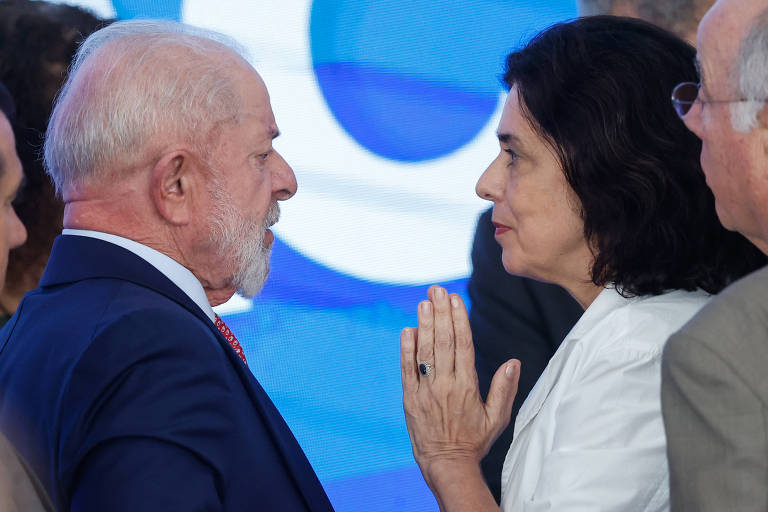 A imagem mostra dois indivíduos em uma conversa próxima. À esquerda， um homem de cabelo grisalho e barba， vestindo um terno escuro e gravata vermelha. À direita， uma mulher com cabelo escuro e liso， usando uma blusa branca. Ela está com as mãos juntas em um gesto que sugere respeito ou solicitação. Ao fundo， há uma tela com formas azuis.