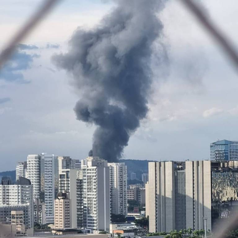 A imagem mostra uma densa coluna de fumaça preta subindo em direção ao céu， localizada em uma área urbana com vários edifícios. A fumaça se destaca contra um céu parcialmente nublado. A vista é em perspectiva， com uma grade visível em primeiro plano， sugerindo que a foto foi tirada de uma janela ou varanda.