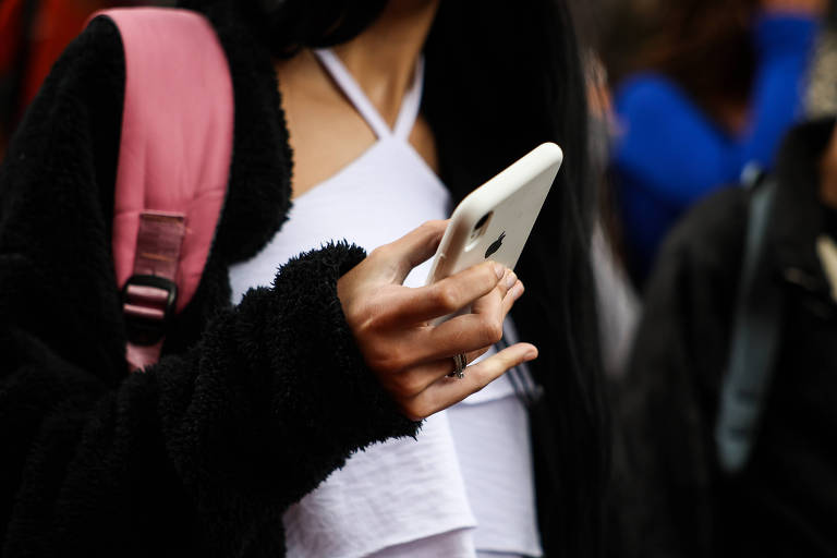 A imagem mostra uma pessoa segurando um celular na mão direita. A pessoa está vestindo uma blusa branca com alças e um casaco preto de pelúcia. Ela também tem uma mochila rosa nas costas. O fundo é desfocado， com outras pessoas visíveis.