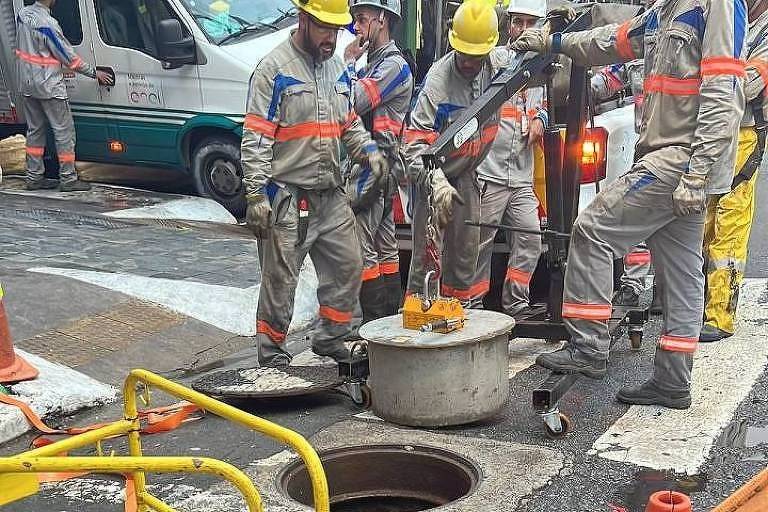 Homens vestidos com uniforme da Enel e equipamentos de segurança retiram， com a ajuda de um guindaste， uma tampa de concreto de uma galeria subterrânea de fiação da concessionária emSão Paulo