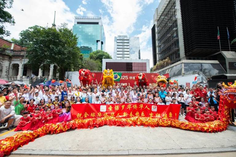 A imagem mostra um grande grupo de pessoas reunidas em uma celebração do Ano Novo Chinês. No centro， há uma faixa vermelha com caracteres chineses. Algumas pessoas estão vestidas com roupas tradicionais， enquanto outras usam camisetas coloridas. Dois leões dançantes estão presentes， um vermelho e um amarelo. Ao fundo， há prédios modernos e árvores， sob um céu azul com algumas nuvens.