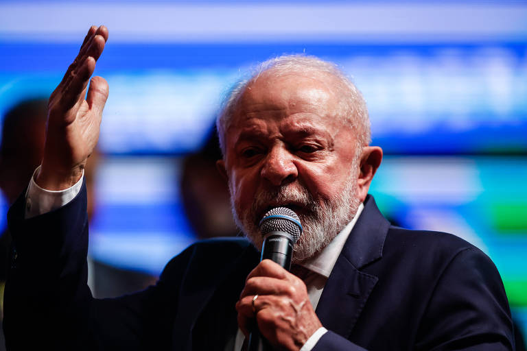 Lula tem cabelo grisalho e barba， veste com um terno escuro e está falando ao microfone. Ele levanta a mão direita em um gesto expressivo， enquanto parece se dirigir a uma audiência. O fundo é desfocado， com luzes coloridas que sugerem um ambiente de evento ou conferência.