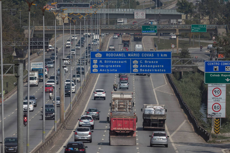 A imagem mostra uma estrada movimentada com vários veículos， incluindo carros e caminhões. Há placas de sinalização em azul indicando direções e destinos， além de sinais de trânsito ao lado da estrada. O ambiente é urbano， com árvores ao fundo