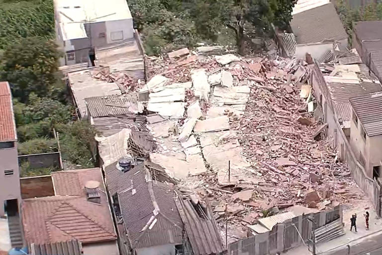 A imagem mostra a área de um desabamento， onde uma estrutura de edifício colapsou， deixando escombros espalhados. Ao redor， há casas e vegetação. Algumas pessoas estão visíveis na cena， observando os destroços.