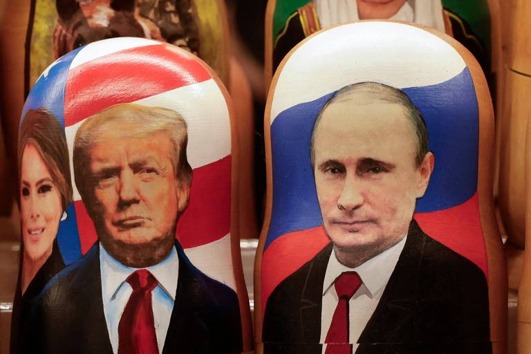 A imagem mostra bonecos matrioshka， também conhecidos como bonecas russas， com retratos de Donald Trump e Vladimir Putin. O boneco à esquerda apresenta a imagem de Trump， com uma bandeira dos Estados Unidos ao fundo， enquanto o boneco à direita exibe Putin， com a bandeira da Rússia. Uma figura feminina， possivelmente Melania Trump， está parcialmente visível ao lado de Trump.