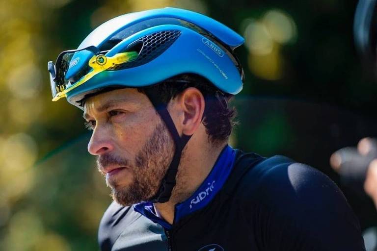 Um ciclista está usando um capacete azul e uma camisa de ciclismo preta com detalhes em azul. Ele parece concentrado e está em um ambiente ao ar livre， com luz natural ao fundo. O ciclista tem barba e cabelo curto.