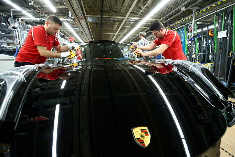 A imagem mostra dois trabalhadores em uma linha de montagem， ambos vestindo camisetas vermelhas， enquanto trabalham em um carro preto. O carro possui um emblema da Porsche visível na parte frontal. O ambiente é uma fábrica， com iluminação fluorescente e equipamentos ao fundo.
