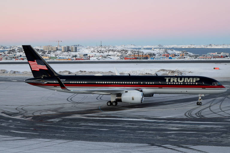 A imagem mostra um avião particular preto com detalhes dourados e vermelhos， estacionado em uma pista coberta de neve. O nome quot;TRUMPquot; está visível na lateral da aeronave， e a cauda apresenta uma bandeira dos Estados Unidos estilizada. Ao fundo， há uma paisagem nevada com algumas construções e luzes acesas， sugerindo um ambiente frio， possivelmente durante o amanhecer ou anoitecer.