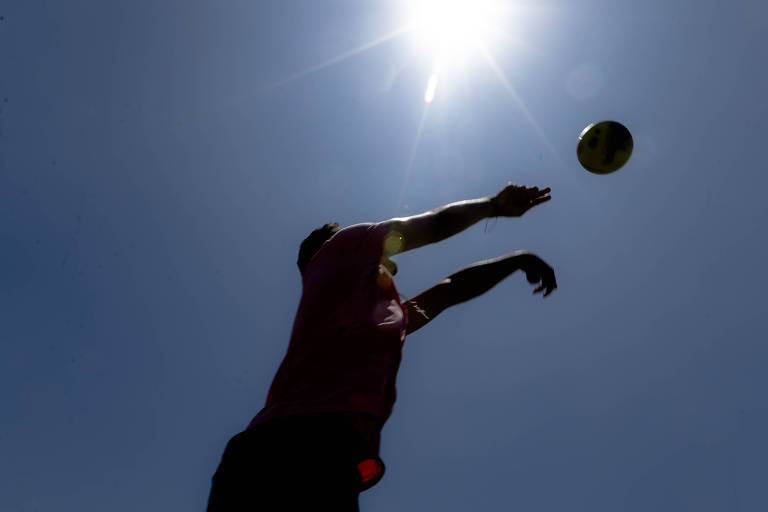 A imagem mostra a silhueta de um jogador de vôlei saltando para fazer um saque. O sol brilha intensamente no céu, criando um contraste forte com a figura do atleta. Ao fundo, é possível ver a vegetação e outras pessoas, mas estão em sombras devido à iluminação.