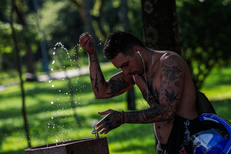 Um homem com tatuagens está em pé ao lado de uma fonte de água em um parque. Ele está sem camisa， usando fones de ouvido e parece estar se preparando para beber água. A água está jorrando da fonte， criando um efeito de gotículas ao redor. O fundo é composto por uma grama verde e árvores.