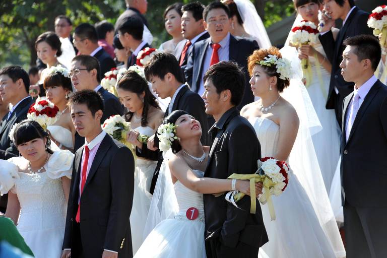 A imagem mostra um grupo de pessoas em um evento de casamento. No centro， um casal de noivos se destaca， com a noiva usando um vestido branco e segurando um buquê de flores. Ao redor deles， há várias pessoas vestidas formalmente， algumas em trajes de noiva e outras em ternos. A maioria das mulheres tem flores nos cabelos e seguram buquês. O ambiente parece ser ao ar livre， com árvores ao fundo.