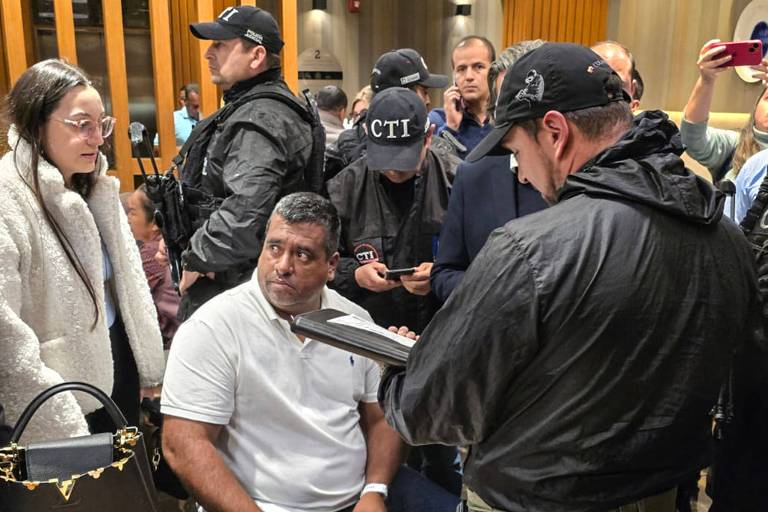 A imagem mostra um grupo de pessoas em um ambiente interno， possivelmente uma sala de reuniões ou um auditório. Um homem está sentado à mesa， com uma expressão séria， enquanto um agente de segurança， vestido de preto， está próximo a ele， segurando um dispositivo. Ao fundo， há várias pessoas observando a cena， algumas com celulares. Uma mulher com óculos e uma jaqueta clara está ao lado do homem sentado. Outros agentes de segurança estão visíveis， todos usando roupas escuras e bonés.