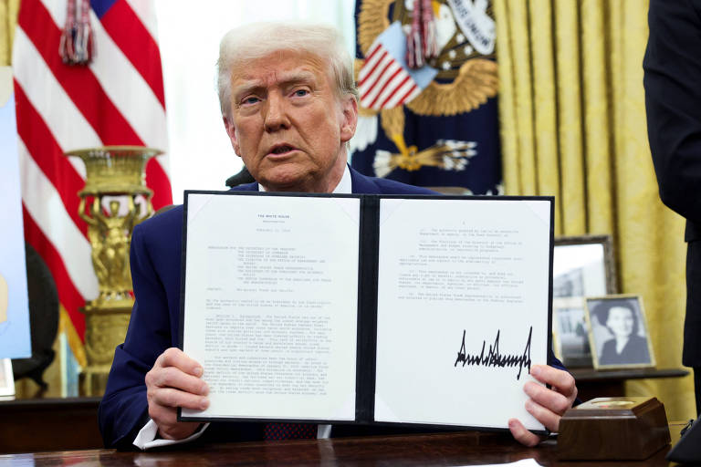 Um homem com cabelo loiro e uma gravata vermelha está sentado em uma mesa， segurando um documento assinado. Ao fundo， há bandeiras dos Estados Unidos e um emblema presidencial. O ambiente é um escritório com cortinas douradas e uma foto emoldurada visível na mesa.