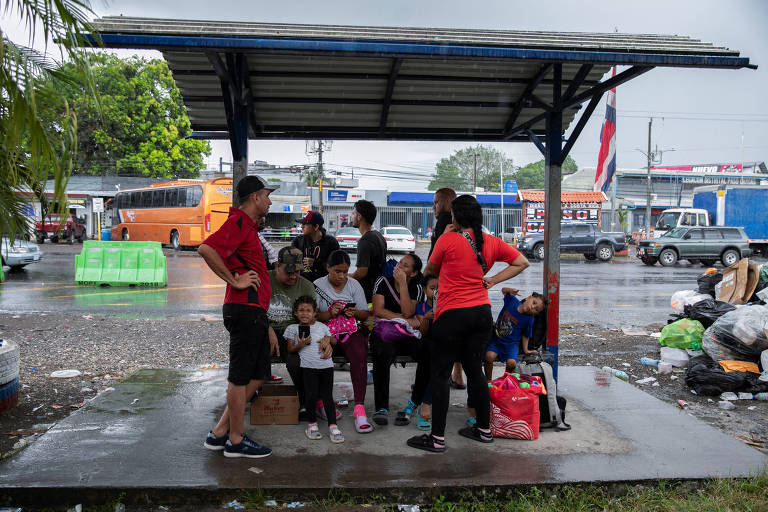 Um grupo de pessoas se abriga sob uma cobertura em uma área urbana durante a chuva. Algumas crianças estão sentadas no chão， enquanto adultos conversam em pé. Ao fundo， há veículos estacionados e lixo acumulado no chão， indicando um ambiente urbano. O céu está nublado e chuvoso.