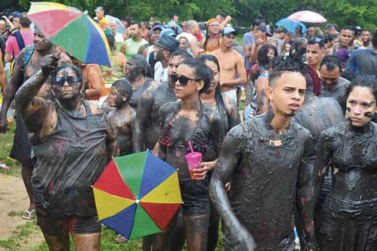 A imagem mostra um grupo de pessoas participando de uma festa ao ar livre， todas cobertas de lama. Algumas pessoas seguram guarda-chuvas coloridos， enquanto outras têm copos na mão. O fundo é composto por uma multidão de pessoas， algumas com roupas de banho e outras com roupas casuais. O ambiente parece festivo， com árvores e um céu nublado.