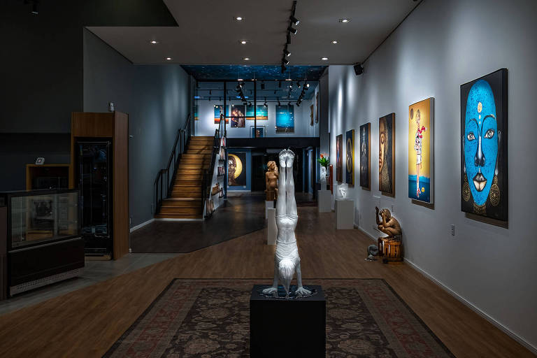 A imagem mostra o interior de uma galeria de arte moderna. No centro, há uma escultura de uma figura humana em posição de cabeça para baixo, em uma base preta. As paredes estão adornadas com várias obras de arte, incluindo retratos e pinturas coloridas. À esquerda, há uma vitrine e uma escada que leva a um andar superior. O piso é de madeira e há um tapete decorativo no chão.