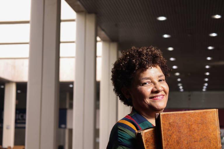 A imagem mostra uma mulher sorridente com cabelo cacheado, segurando livros em suas mãos. Ela está em um ambiente interno com paredes claras e iluminação natural. O fundo apresenta colunas e um teto com iluminação artificial.