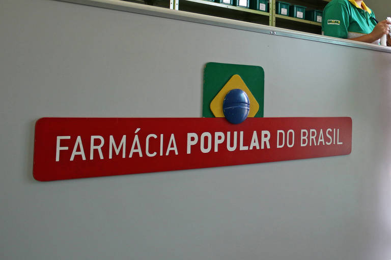 A imagem mostra o interior de uma farmácia popular no Brasil. No balcão, uma mulher está conversando com duas funcionárias que usam uniformes verdes. Ao fundo, há prateleiras com caixas organizadas. Na parede, há um letreiro vermelho com o texto 'FARMÁCIA POPULAR DO BRASIL' e um símbolo da bandeira do Brasil.