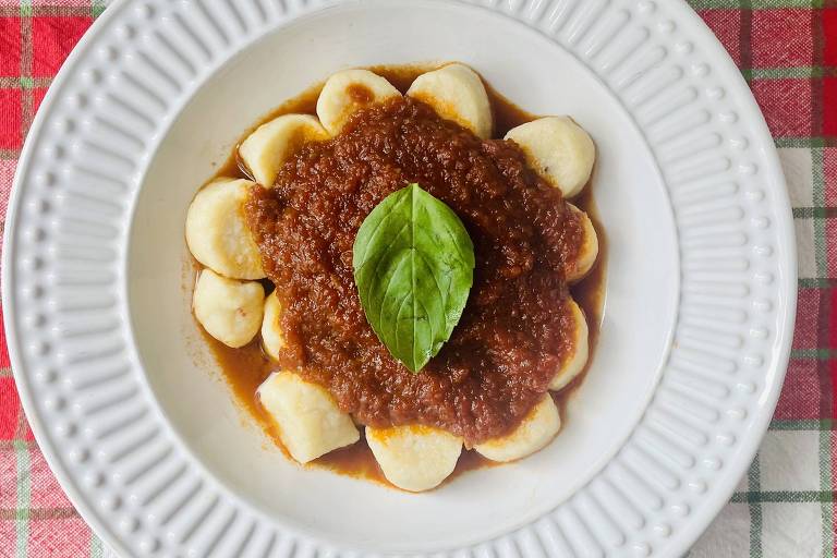 Um prato de gnocchi servido em um prato branco. O gnocchi é disposto em forma de flor ao redor de um molho marrom espesso， que está no centro. Uma folha de manjericão verde está colocada sobre o molho. O fundo é uma toalha de mesa quadriculada em vermelho e branco.