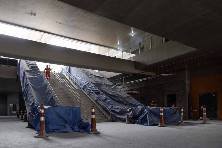 A imagem mostra um espaço interno de um edifício em construção， com duas escadas rolantes cobertas por lonas azuis. Há um trabalhador vestido com um macacão laranja em cima de uma das escadas. O ambiente é amplo， com paredes de concreto e iluminação artificial visível no teto.