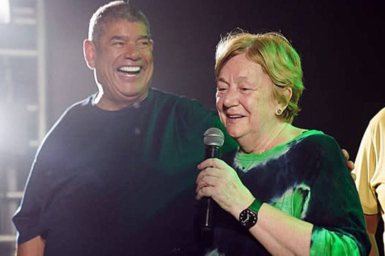 A imagem mostra um homem e uma mulher sorrindo em um evento. O homem， à esquerda， está usando uma camiseta escura e segurando um microfone， enquanto a mulher， à direita， está com uma blusa colorida e parece emocionada. Ambos estão em um ambiente iluminado， com luzes ao fundo.