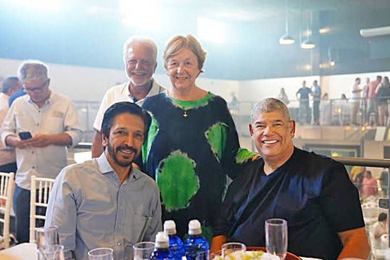 A imagem mostra um grupo de quatro pessoas em um evento social. Dois homens estão sentados à mesa， um deles com cabelo escuro e barba， vestindo uma camisa clara， e o outro com cabelo curto e uma camiseta preta. Atrás deles， há um casal de pessoas mais velhas， um homem com cabelo grisalho e uma mulher com cabelo curto e uma blusa verde. O ambiente é iluminado e parece ser um espaço de evento com mesas e cadeiras brancas.