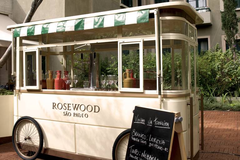 A imagem mostra um carrinho de bebidas localizado no Rosewood São Paulo. O carrinho é de cor clara, com uma cobertura listrada em verde e branco. Na parte frontal, está escrito 'ROSEWOOD SÃO PAULO'. Ao lado do carrinho, há uma placa de madeira preta com informações sobre os vinhos disponíveis, incluindo 'Vinho na Torneira', 'Branco: Lorena', 'Tinto: Nebbiolo' e o preço de R$ 39. Ao fundo, pode-se ver um edifício com janelas verdes e uma vegetação ao redor.