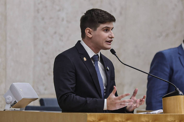 Um jovem homem， vestido com um terno escuro e gravata， está falando em um microfone durante uma sessão legislativa. Ele gesticula com as mãos enquanto se expressa. Ao fundo， há uma mesa e outras pessoas， mas não são claramente visíveis.