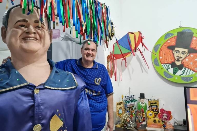 A imagem mostra um ambiente decorado para uma festa， com um boneco grande sorridente à esquerda， vestindo uma roupa azul. À direita， uma pessoa está sorrindo e posando para a foto. O fundo é decorado com fitas coloridas penduradas e um quadro circular com uma ilustração de um homem de chapéu. Há também várias peças 