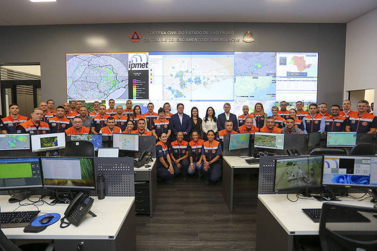 A imagem mostra um grupo grande de pessoas em um centro de comando， posando para a foto. Elas estão vestidas com uniformes laranja e azul. Ao fundo， há várias telas exibindo mapas e dados. O ambiente é moderno， com mesas e computadores visíveis na parte inferior da imagem.