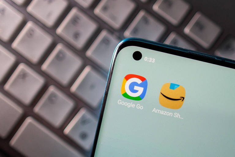 A imagem mostra a tela de um smartphone com dois ícones de aplicativos visíveis: o ícone do Google Go， que é um 039;G039; colorido， e o ícone do Amazon Shopping， que é um símbolo de carrinho de compras. Ao fundo， é possível ver um teclado de computador desfocado.