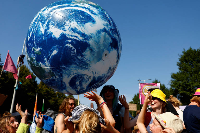 A imagem mostra um grupo de pessoas em uma manifestação ao ar livre， com um grande globo terrestre inflável sendo jogado para o alto. Algumas pessoas estão sorrindo e tentando pegar o globo， enquanto outras observam. O céu está claro e ensolarado， e há bandeiras e cartazes ao fundo.