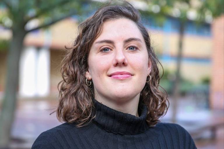 A imagem mostra uma mulher com cabelo cacheado e castanho claro， usando um suéter preto de gola alta. Ela está sorrindo levemente e posando em um ambiente ao ar livre， com árvores e edifícios ao fundo. A iluminação é natural， sugerindo um dia claro.