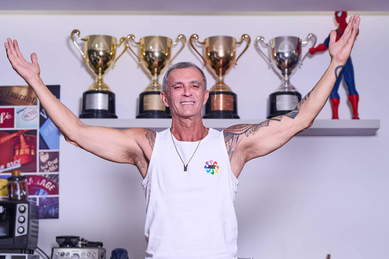 Um homem de camiseta branca levanta os braços em um ambiente interno. Ao fundo, há uma prateleira com vários troféus dourados e prateados. O homem tem cabelo curto e grisalho, e está sorrindo. Na parede, há uma colagem de imagens e objetos decorativos, incluindo uma figura do Homem-Aranha.