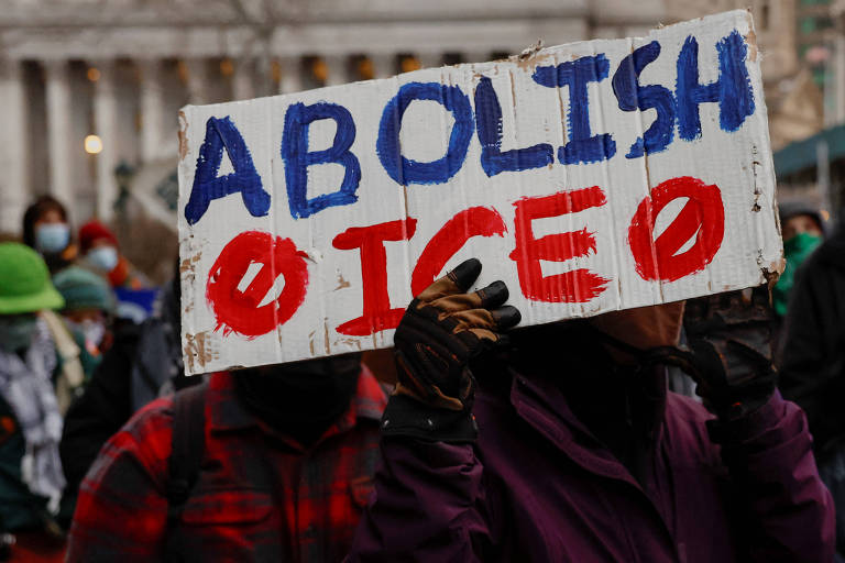 Uma pessoa segurando um cartaz que diz 039;ABOLISH ICE039; em letras grandes e coloridas. O fundo mostra um grupo de pessoas， algumas usando máscaras， em um ambiente urbano. A atmosfera é de protesto， com pessoas vestindo roupas de inverno.