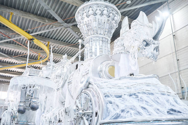A imagem mostra um carro alegórico predominantemente branco, com detalhes em prata. O carro possui uma estrutura elaborada, incluindo uma figura de um dragão na parte frontal e elementos decorativos que lembram uma carruagem. O ambiente ao redor é um galpão com teto alto e estrutura metálica visível.